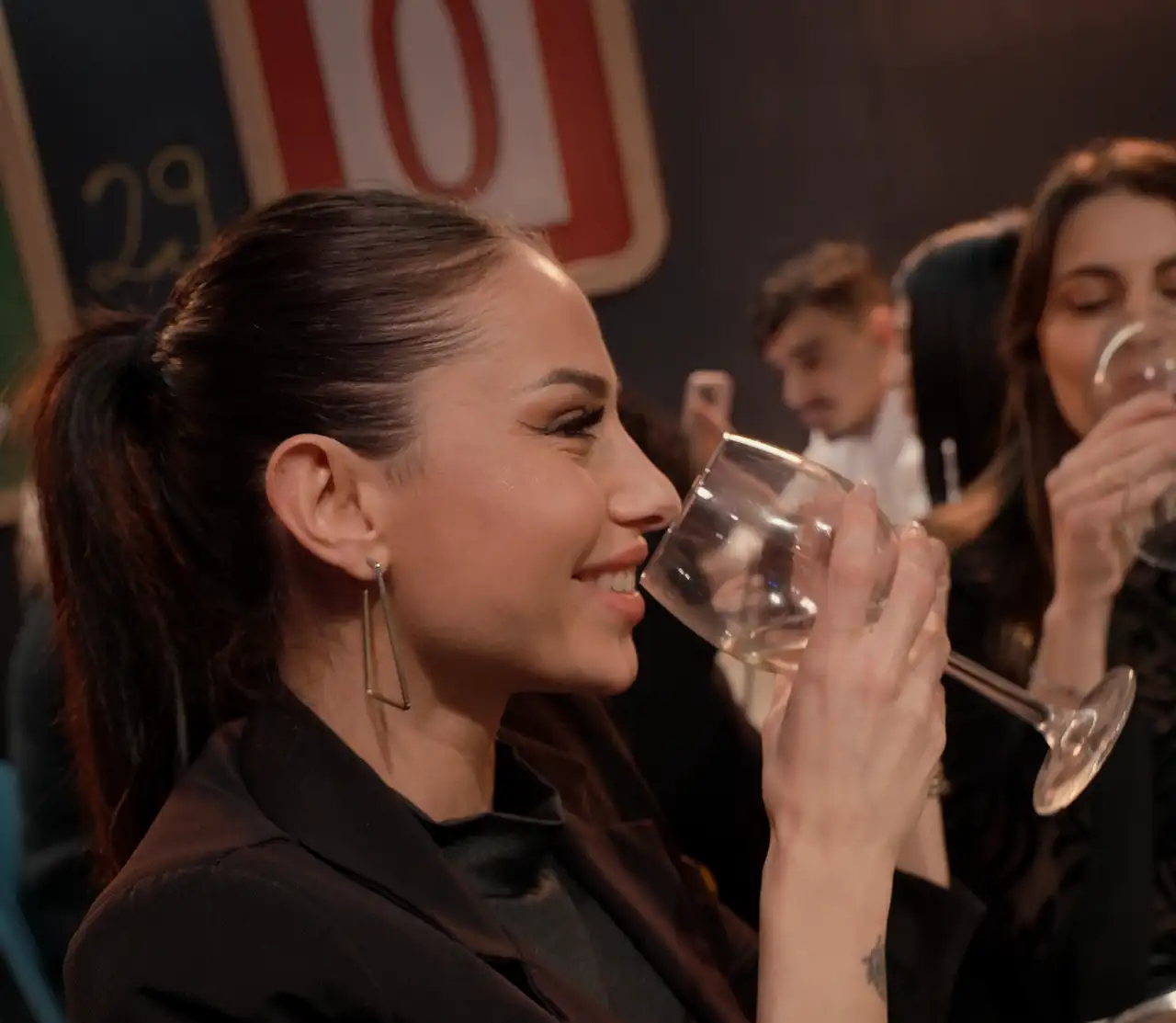 Una ragazza beve un calice di vino bianco al tavolo, la sala piena alle sue spalle